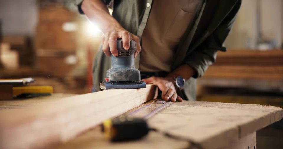 Photographie en atelier de menuiserie: deux menuisiers de générations différentes (un plus âgé, un plus jeune) travaillant ensemble sur une pièce de bois, l'aîné guidant ou consultant avec le plus jeune. Environnement d'atelier authentique avec machines, établis, copeaux de bois, lumière naturelle. Moment de transmission du savoir-faire et de collaboration familiale intergénérationnelle.